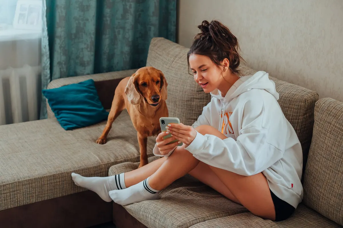 A woman sits on a sofa with her dog beside her, looking at her smartphone.