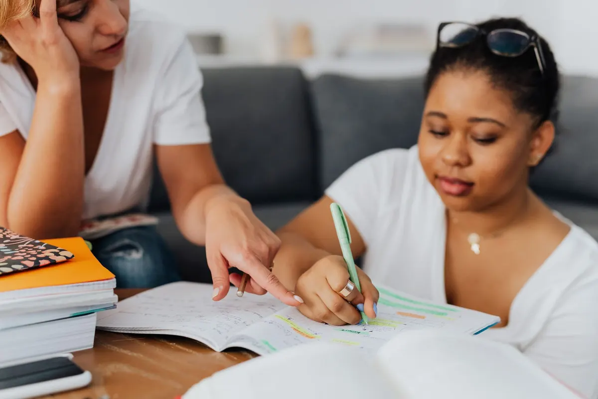 A tutor sits beside a teenage student at a table, pointing to a worked maths problem in an exercise book.