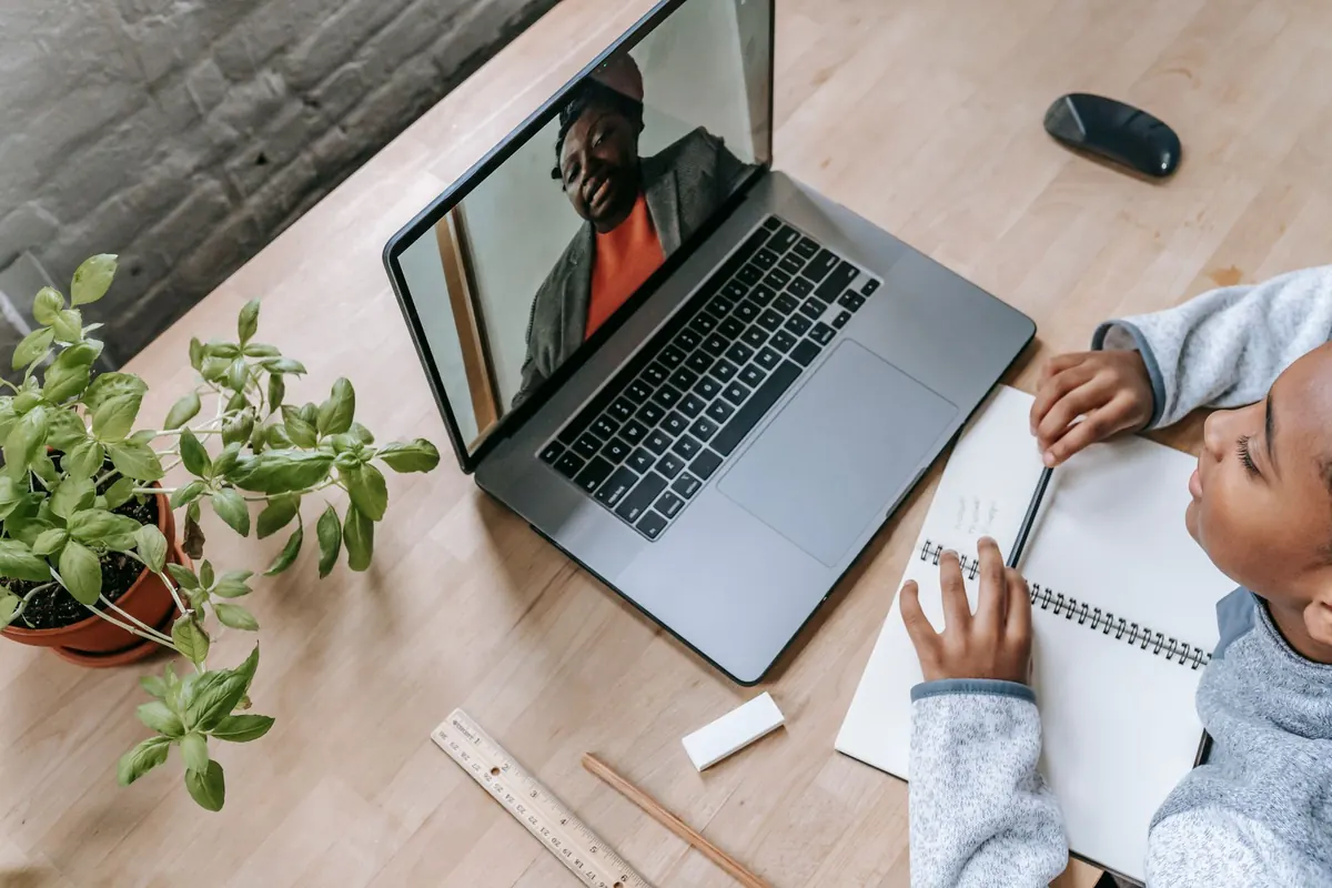 A tutor teaches a lesson over a video call on a laptop, with a notebook and pen beside the keyboard.
