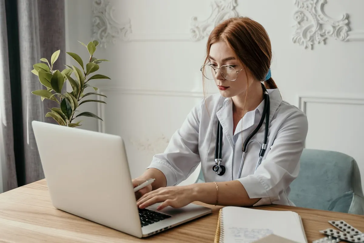 A therapist sits at a wooden desk with a laptop and notebook, writing the text for a practice website.