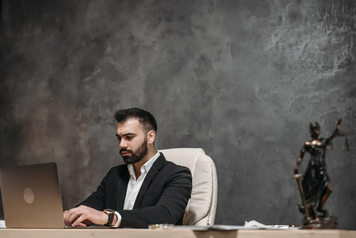 A lawyer works on a laptop at a wooden desk with law books and a notepad beside it.