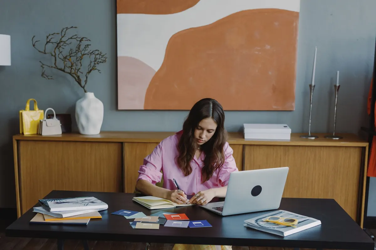 A designer at a desk adjusting a website layout on a laptop beside a notebook of sketches.