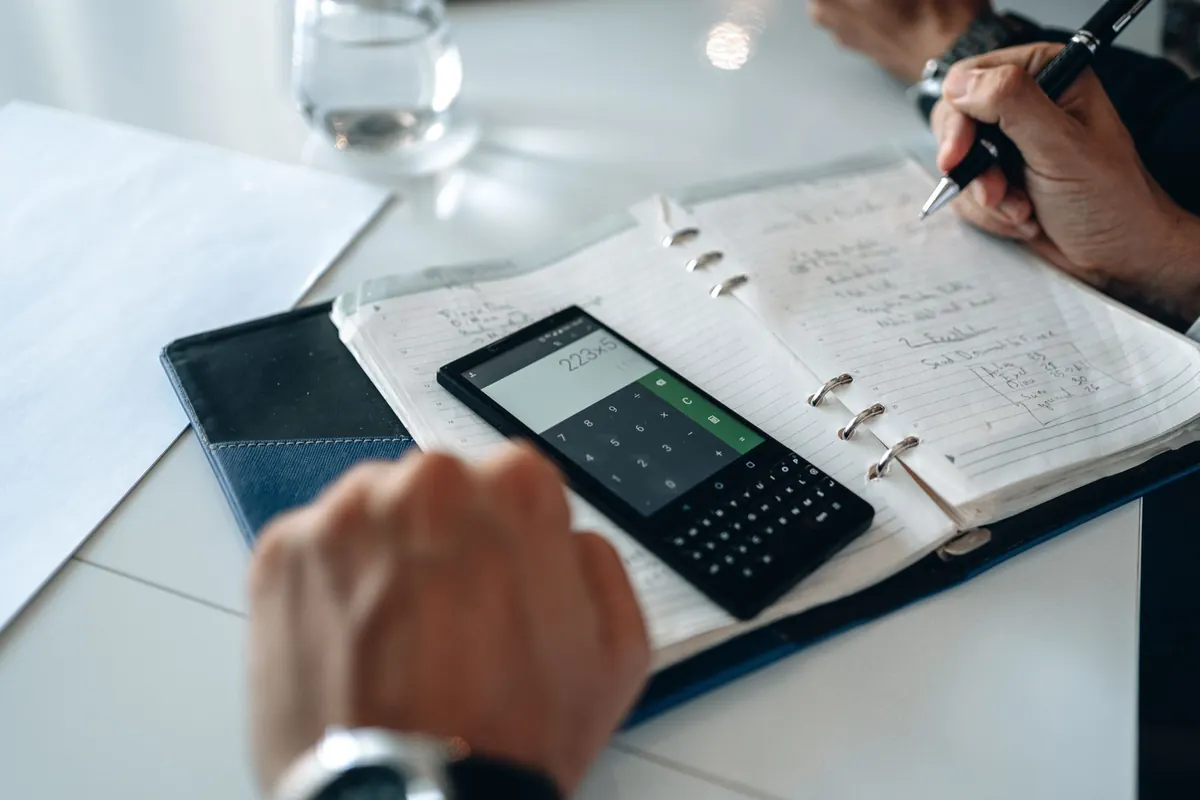 A business owner adds up monthly costs with a calculator and a notebook at a wooden desk.