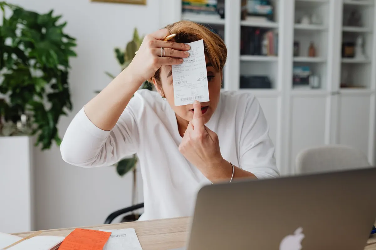 A freelancer at a home office desk checks invoices on a laptop next to a notebook and a cup of coffee.