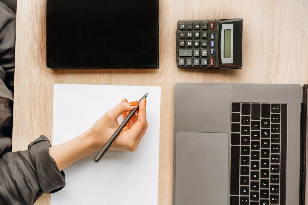 A woman at a desk works through figures on a calculator next to her open laptop.