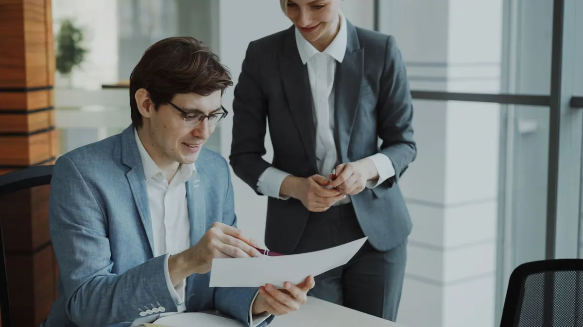 Two colleagues at an office table compare notes and monthly costs on paper and a laptop.