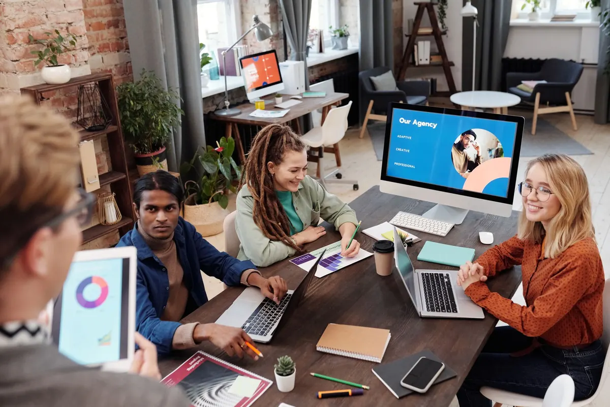 Three people at a small agency sit around a table with laptops, planning work for their clients.