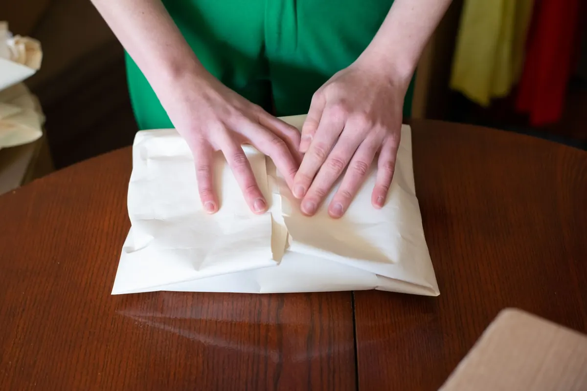 A maker wrapping a small handmade item in tissue paper at a home workbench surrounded by packing supplies.