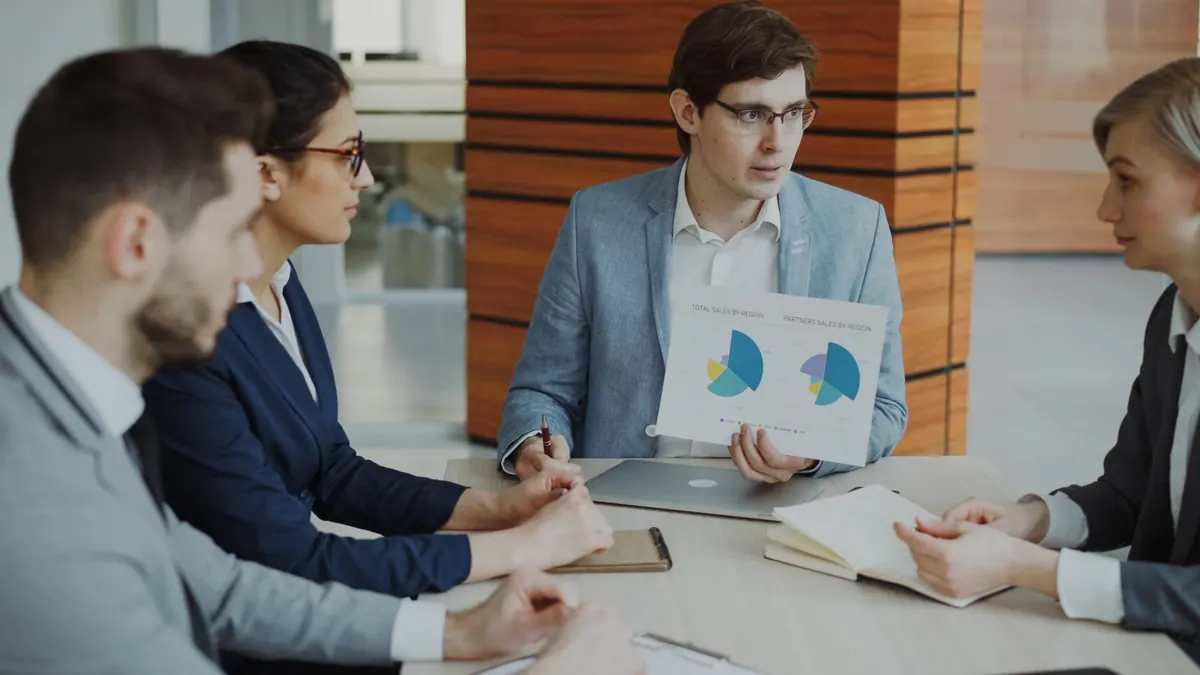A small sales team looks through a list of customer deals together on a laptop in a bright office.