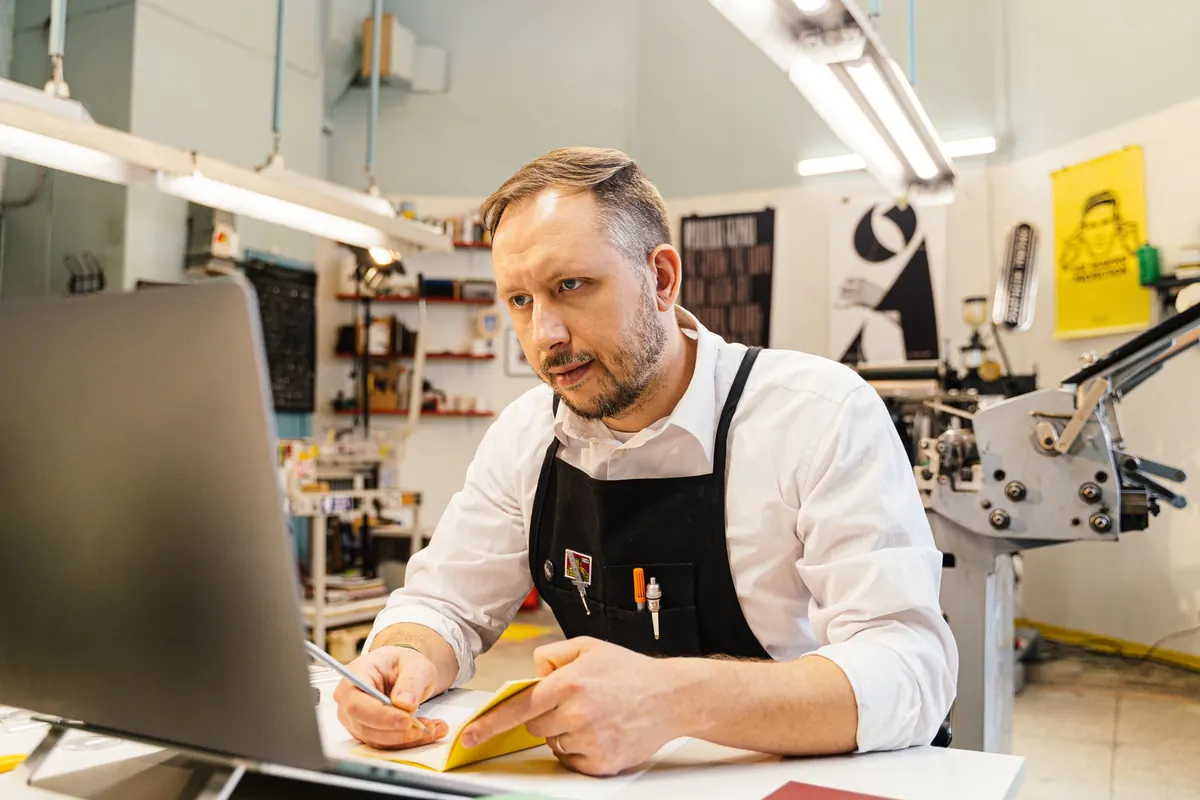 A small shop owner checks orders and customer messages on a laptop beside the shop counter.