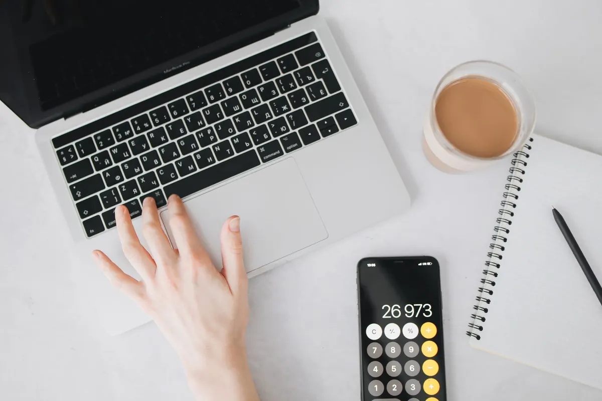 A freelancer works through monthly figures with a calculator and notebook beside an open laptop.