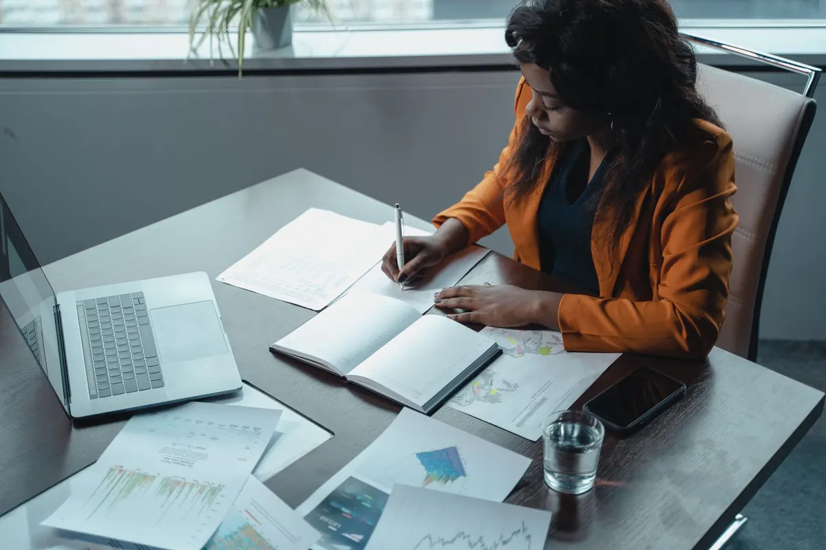 A freelancer at a desk working through a written checklist beside an open laptop.
