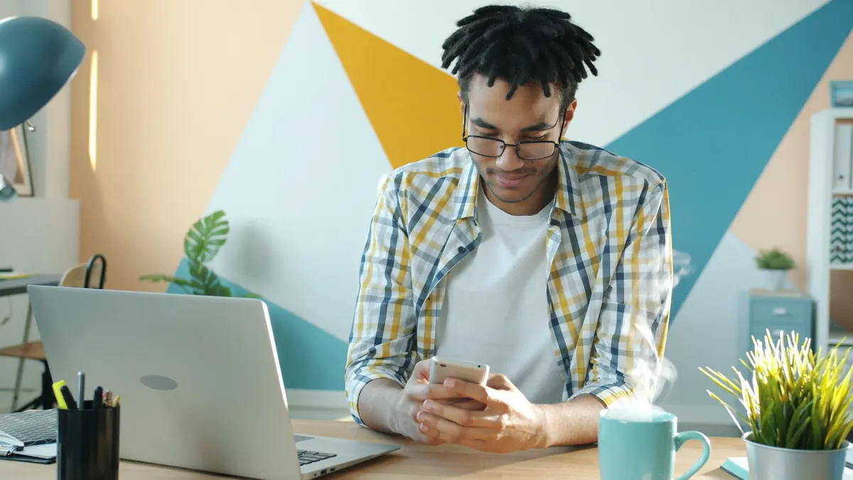 A designer at a desk checks a website on a mobile phone beside an open laptop.