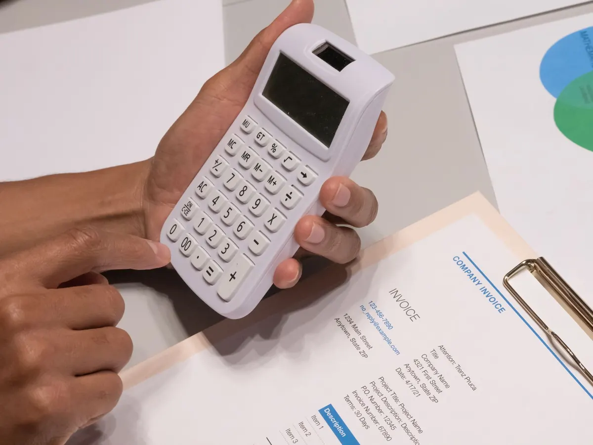 A calculator, a pen and a stack of printed invoices on an office desk.