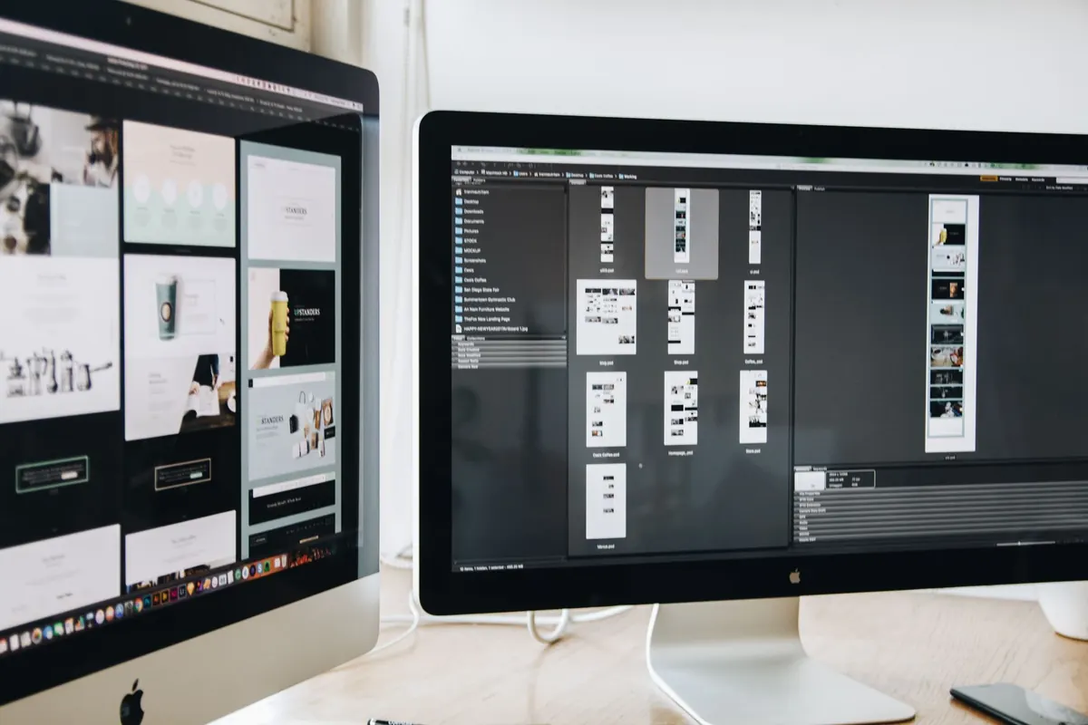 A web designer at a desk with two monitors showing a website layout in progress.