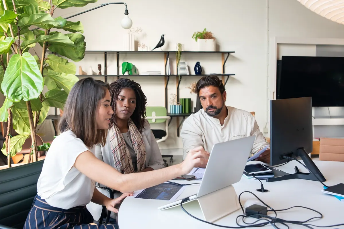 Three colleagues sit around a table in a small office, talking about something on a laptop.